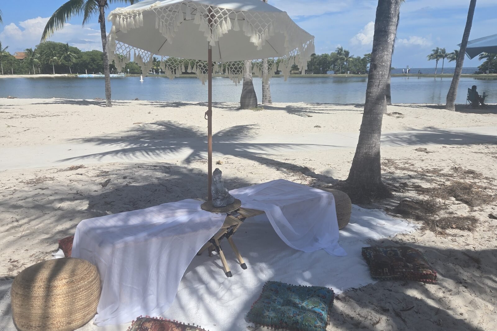 A massage table draped in white under palm trees on the beach, with a parasol, floor cushions and a woven pouffe.